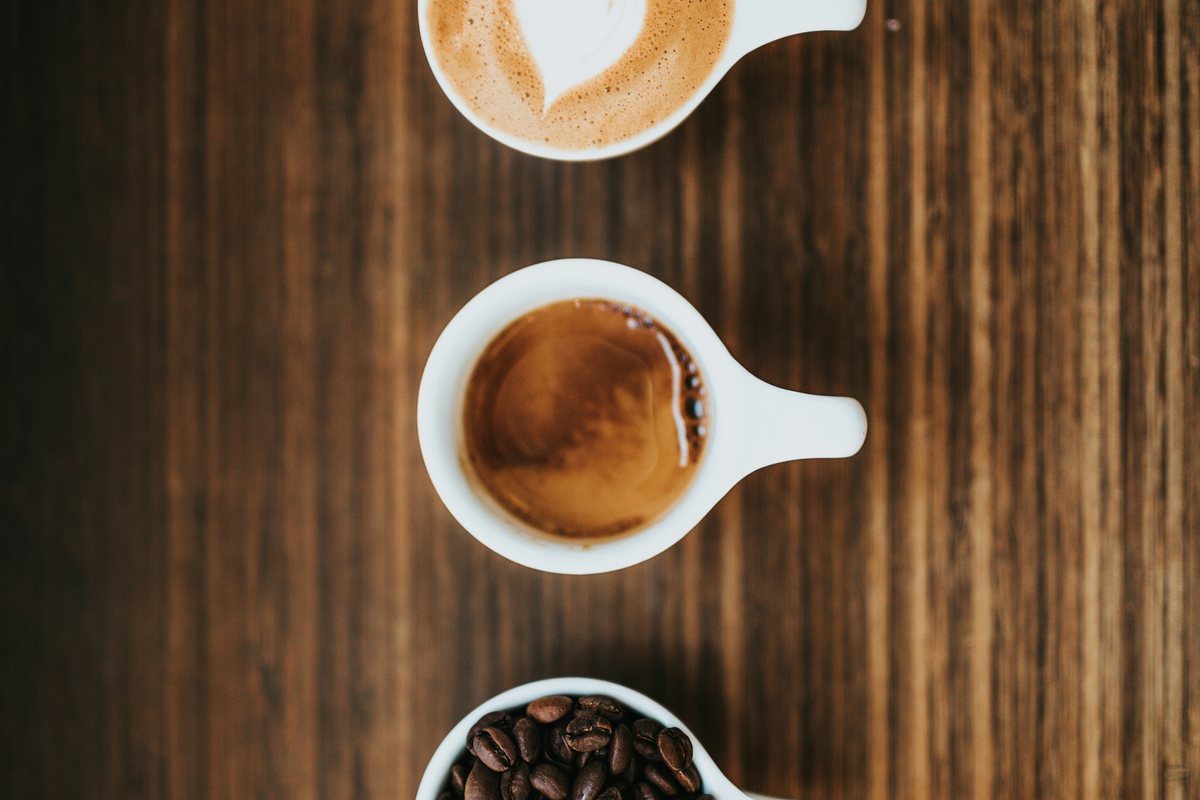 Tři šálky na tmavém dřevě shora: cappuccino, espresso a pražená zrnka.